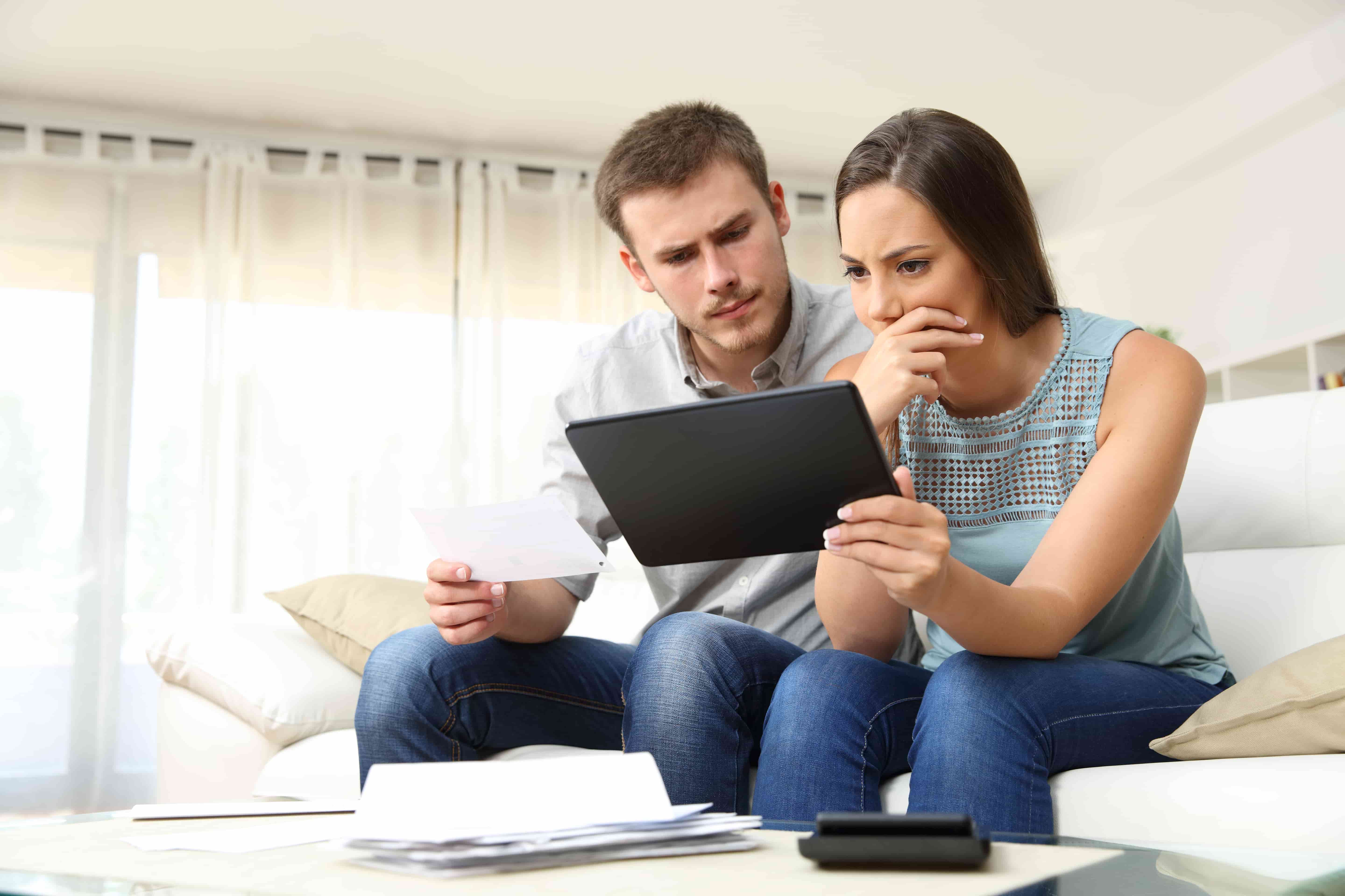 Worried couple checking bank account trouble online in a tablet sitting on a couch in the living room at home